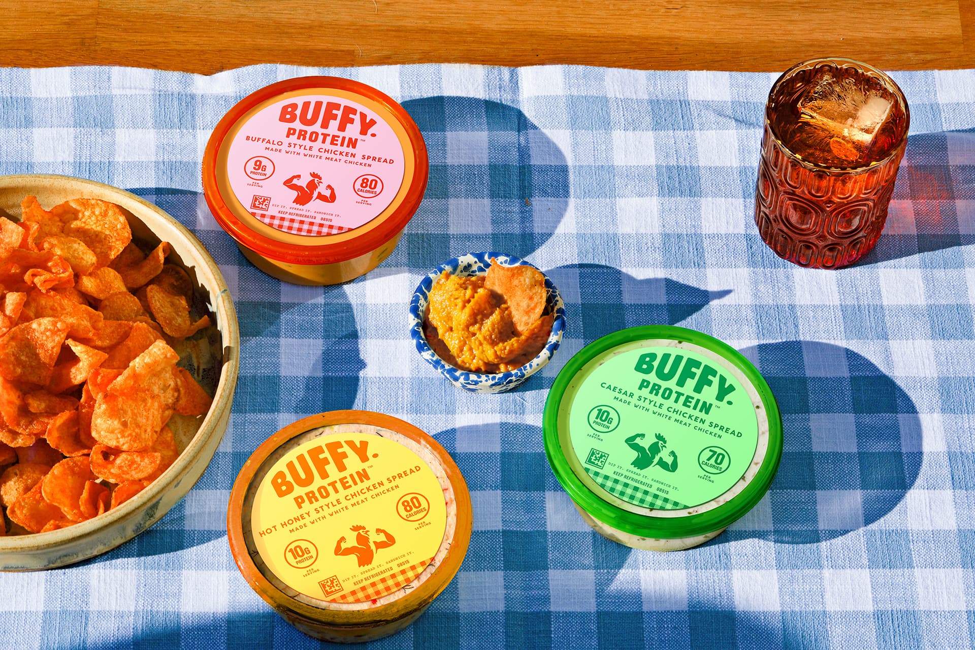Buffy Protein containers on a blue checkered tablecloth with a bowl of chips and a glass of tea.