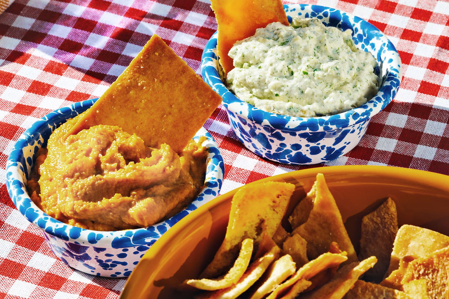 Two bowls of Buffy Protein Dip, one filled with Whoa, Buffalo and the other with Great Caesar. They are sitting on a checkerboard tablecloth with a bowl of pita chips.