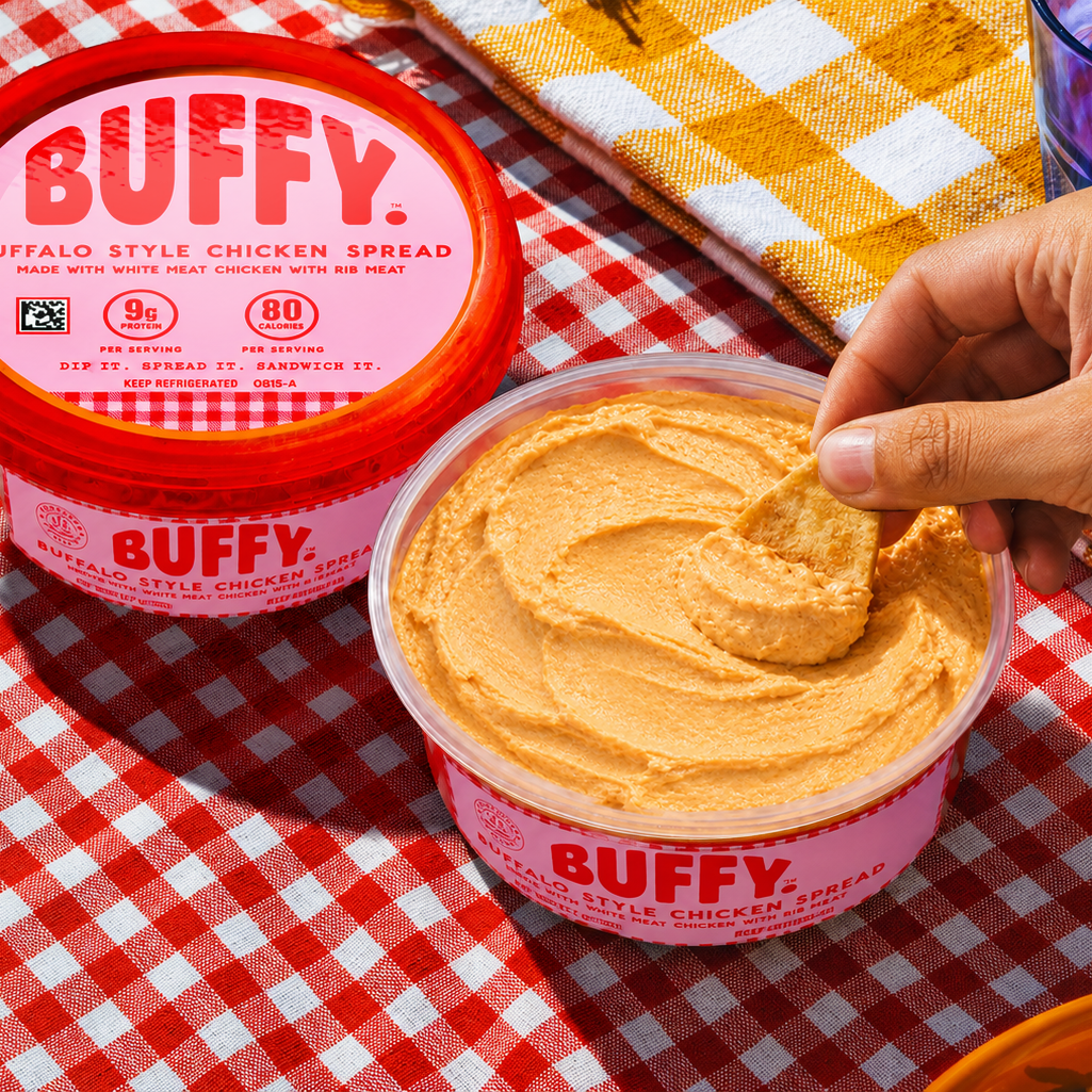 A hand dips a chip into Buffy Protein Buffalo Style Chicken Spread on a red-and-white checkered tablecloth.