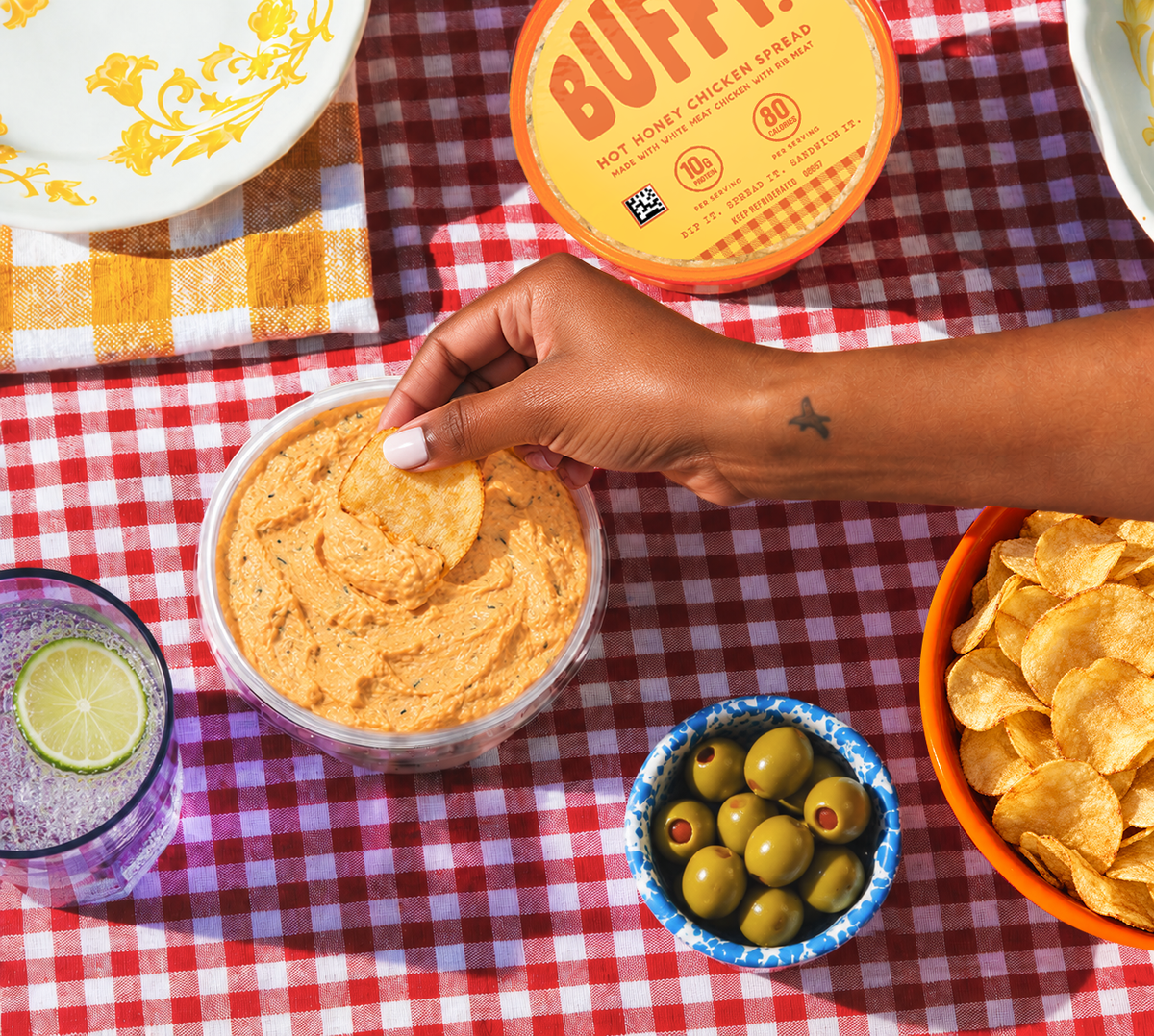 A hand dips a chip into Buffy Protein Hot Honey Chicken Spread on a red-and-white checkered tablecloth, with olives, chips, and a drink.
