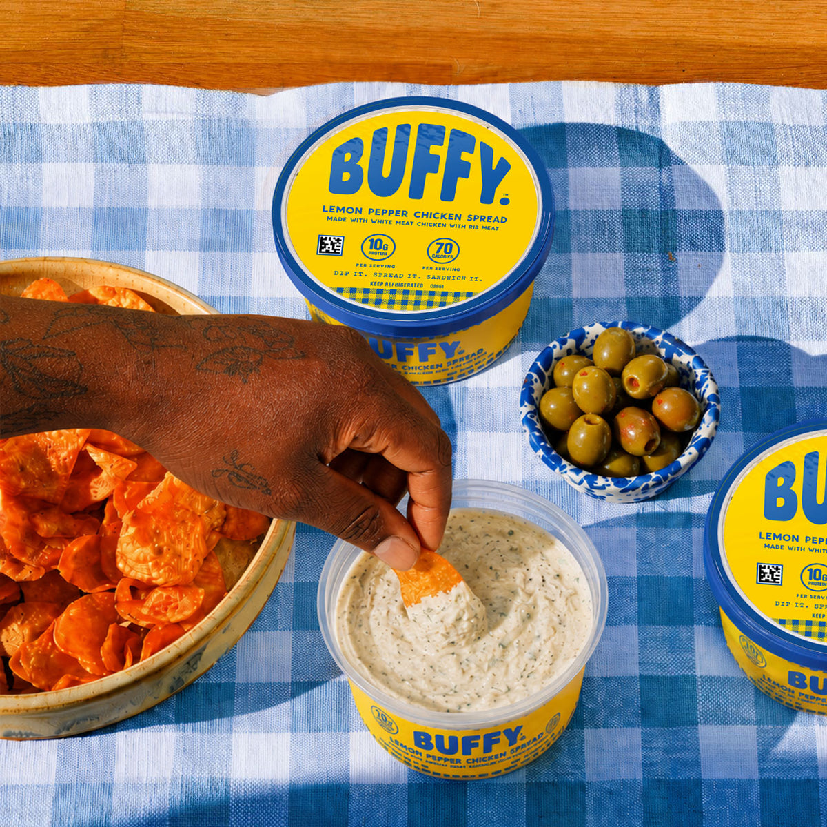 A hand dips a chip into Buffy Protein Lemon Pepper Chicken Spread on a blue-and-white checkered tablecloth, with olives and a bowl of chips.